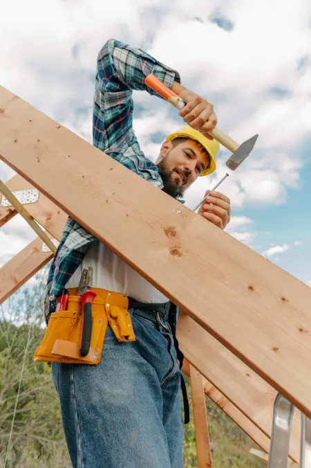 Bauarbeiter mit gelbem Helm schlägt einen Nagel in ein Holzstück, während er auf dem Dach eines Hauses arbeitet.