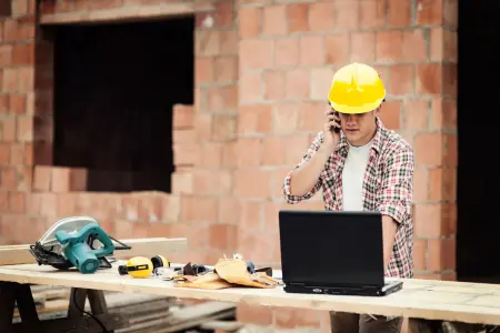 Ein Bauarbeiter mit Schutzhelm telefoniert und arbeitet an einem Laptop auf einer Baustelle.