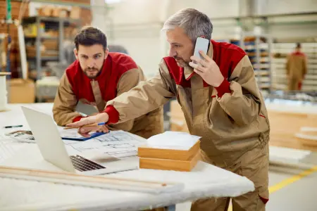 Zwei Arbeiter in einer Werkstatt arbeiten mit einem Laptop und besprechen Baupläne.